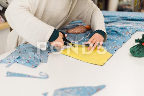 Unrecognizable female tailor in casual sweater cutting piece of colorful fabric with scissors while working on project in creative atelier {{{woman,fabric,cut,tailor,atelier,dressmaker,scissors,designer,creative,textile,small business,work,piece,seamstress,job,workshop,design,skill,occupation,female,craft,handmade,handicraft,fashion,cloth,material,workplace,sew,hobby,studio,table,busy,create,sewer,desk,master,workspace,tool,process,parlor,pattern,handwork,manufacture,manual,industry,unrecognizable,faceless,anonymous,crop}}}