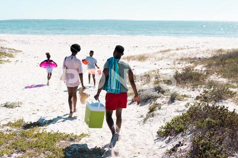 Rear view of african american family with swimming float, cooler walking at beach towards seascape. Copy space, unaltered, parents, together, childhood, picnic, nature, vacation, enjoyment, summer. Four People, Mid Adult, Man, Male, Black, Woman, Female, Child, Girl, Boy, People, Rear View, African American, Family, Swimming, Float, Cooler, Walking, Beach, Towards, Seascape, Copy Space, Unaltered, Parent, Together, Childhood, Picnic, Nature, Kid, Mother, Sea, Ocean, Little, Daughter, Coast, Summer, Vacation, Sand, Water, Mom, Fun, Happiness, Father, Son, Happy, Outdoor, Shore, Love, Rear, View, African, American, Copy, Space