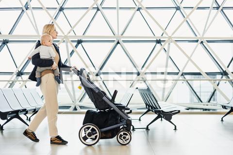 Mother carying his infant baby boy child, pushing stroller at airport departure terminal moving to boarding gates to board an airplane. Family travel with baby concept travel, family, mother, airport, flight, departure, kid, trip, passenger, boy, journey, tourist, transportation, tourism, stroller, pushing, vacation, plane, terminal, woman, toddler, airplane, lifestyle, parent, infant, mom, transport, childhood, girl, gate, destination, gates, children, baggage, airline, voyage, cart, window, tour, boarding, carry, moving home, wait, cary, board, aerport, station