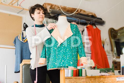 Concentrated female tailor in casual clothes with measuring tape on neck standing near mannequin while preparing clothing for work in creative studio {{{woman,tape,mannequin,studio,workshop,tailor,dressmaker,measure,atelier,designer,work,cloth,female,creative,concentrate,seamstress,professional,craft,occupation,focus,handicraft,create,prepare,fashion,skill,style,process,talent,handmade,job,workplace,design,busy,stand,small business,handwork,centimeter,apparel,craftswoman,profession,garment,manufacture,serious,textile,master,workspace,fabric,material,blurred background}}}