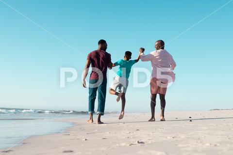 Rear view of african american father and grandfather holding boy's hands and picking him up at shore. Copy space, unaltered, multigeneration family, together, child, nature, vacation, enjoy, summer. Three People, Senior, Elderly, Man, Male, Black, Mid Adult, Child, Boy, People, Rear View, African American, Father, Grandfather, Holding, Hand, Picking, Shore, Copy Space, Unaltered, Multigeneration, Family, Together, Nature, Vacation, Summer, Beach, Mother, Sea, Coast, Parent, Happy, Beautiful, Ocean, Outdoor, Travel, Water, Daughter, Son, Lifestyle, Young, Landscape, Love, Holiday, Rear, View, African, American, Copy, Space