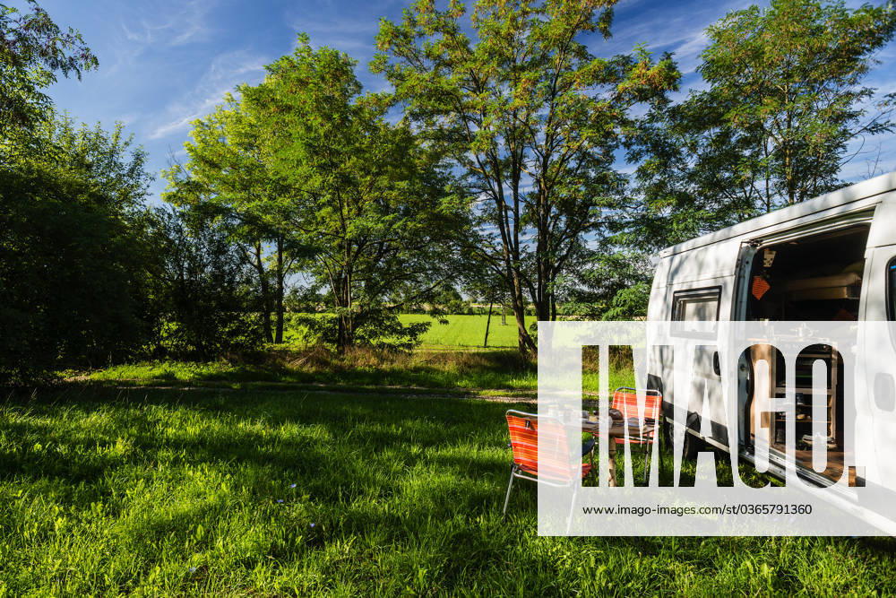 A camper stands between trees on a sunny summers day on a meadow in the countryside, a laid breakfast table with chairs stands in front of the sliding door of the camper Regensburg, Bavaria, Germany In the old town of Regensburg