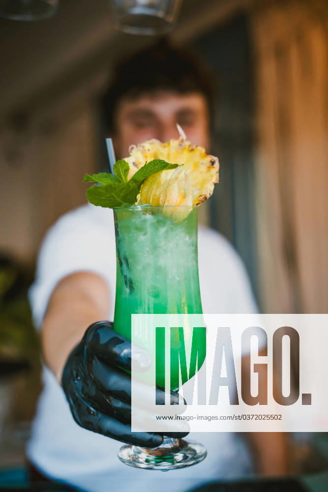 Blurred young male bartender showing a tropical green cocktail with a pineapple garnish at a bar Model Released AlvaroMontero_ID17423_529018_002
