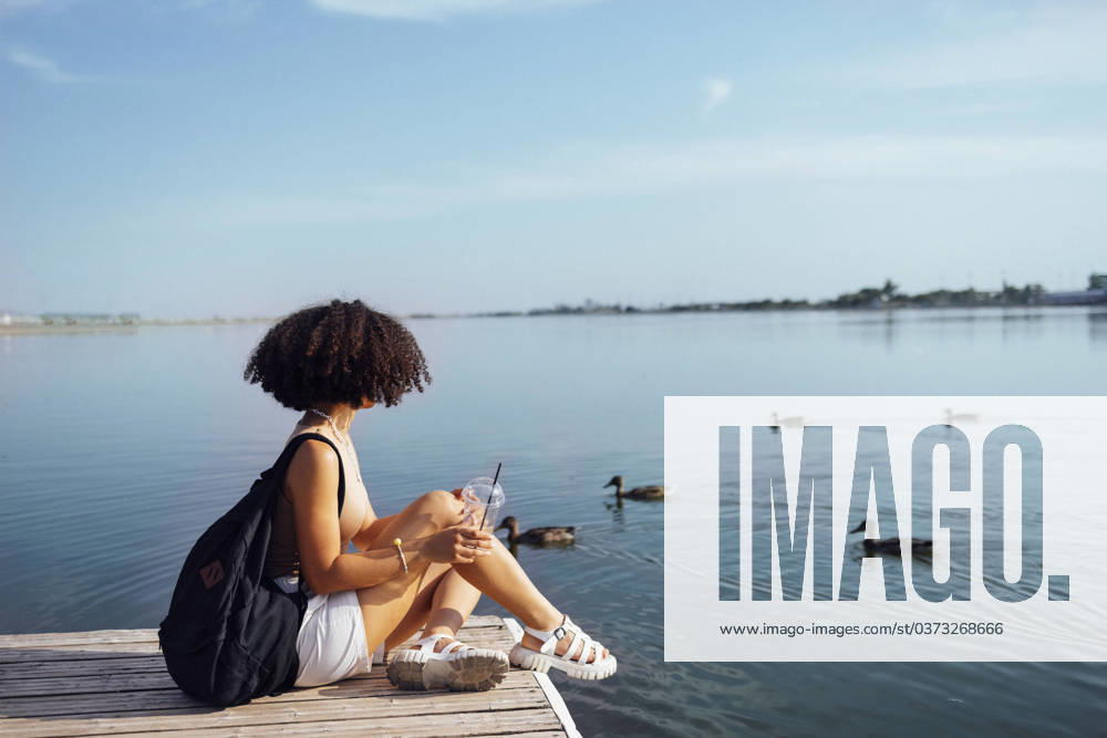 Beautiful mixed race female teenager sits on wooden pier and drinks cocktail in plactic glass. Charming african girl in casual clothes and with black backpack smiles sincerely near water. Sunny day. Beautiful mixed race female teenager sits on wooden pier and drinks cocktail in plactic glass. Charming african girl in casual clothes and with black backpack smiles sincerely near water. Sunny day. ,model released, Symbolfoto