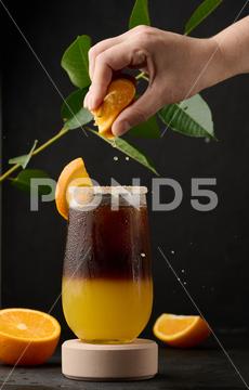 Iced coffee with orange juice in a transparent glass, a woman's hand squeezes an orange slice into a bubble drink layer coffee orange brown sweet cool ice espresso cold drink glass fresh juice beverage cocktail morning caffeine fruit syrup table yellow citrus juicy bumble slice nobody black hand drop squeeze