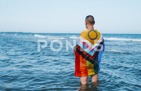 A wet person in the sea wrapped in an intersex-inclusive progress pride flag standing in the water, as seen from behind intersex inclusive progress gay pride flag person man symbol rainbow LGBT LGBTI LGBTQ LGBTIQ homosexual homo homosexuality lesbian transgender transgenderism trans transsexual transsexualism intersexual bisexual bisexuality gender identity orientation queer genderqueer non-binary non binary enby diversity activism activist rights same sex sexual sexuality freedom wet sea ocean outdoors summer horizontal