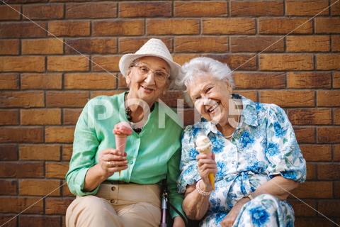 Portrait of two senior female friends in the city, eating ice cream on a hot summer day. Elderly ladies on summer vacation in the city. Pensioners on group trip. Concept of senior vacation and travel groups, clubs. senior city ice cream woman retirement pensioner lifestyle active outdoor elderly old joy vitality happiness smiling eating street togetherness trendy happy holding tourist cone community bonding enjoyment lady older brick wall sitting travel group travel club senior travel traveling vacation chronic illness diabetes heart disease alzheimers disease mental health osteoporosis arthritis dementia hypertension cholesterol depression rheumatoid arthritis incontinence