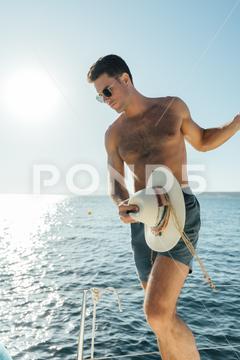 Young fit shirtless man in swimming shorts and hat standing on sailboat enjoying summer vacation against sea and cloudless blue sky {man sailboat yacht boat sensual summer vacation confident sensuality cruise ocean body dreamer stylish vessel model outdoors male natural fit shirtless handsome tourism sunglasses hat transport holidays journey adventure summertime looking away tourist nature sea lifestyle ship style blurred background admire traveler water trip scenic voyage travel relax cloudless peaceful}