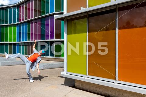 Joyful man dancing energetically in front of a multicolored windowed building {{dancing joyful colorful facade energetic sunglasses daytime street architecture urban city motion style male building reflection modern contemporary outdoors expression vibrant window pane fun happiness dynamic footwear hat playful summer shadow pavement leap design gradient spectrum wall Copy Space}}