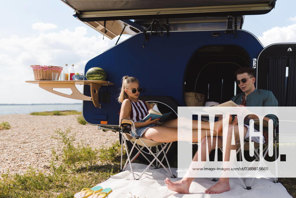 Young couple sitting together in front of van, camping and reading book. Happy young couple sitting together in front of van, camping and reading a book. model released