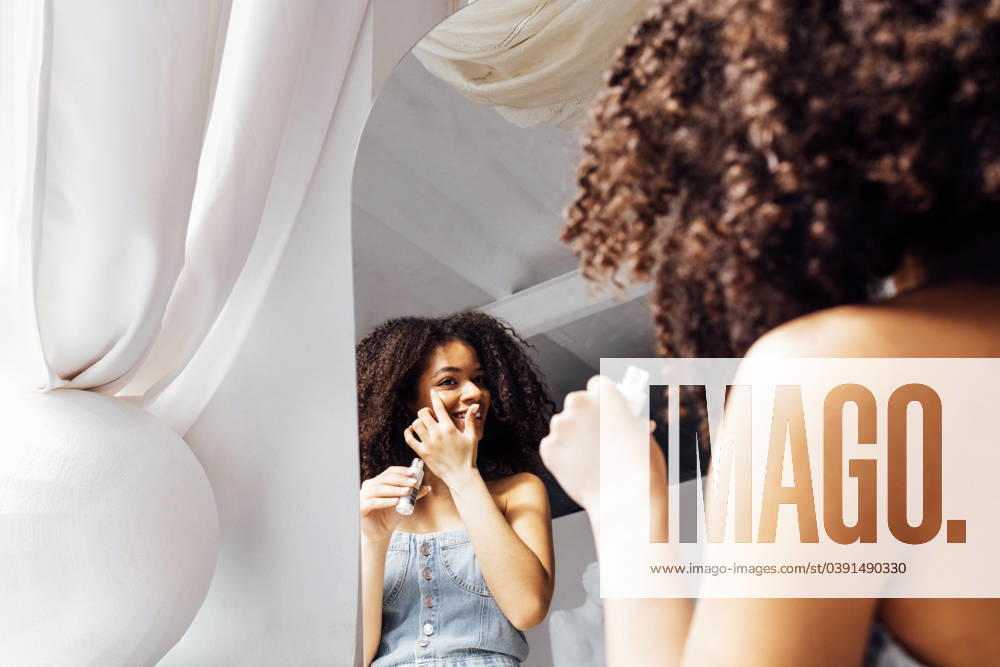 Young pretty african woman in denim top applies glitter gel under her eyes. Young pretty african woman in denim top applies glitter gel under her eyes. Attractive smiling mixed race girl looks in mirror and finishes her makeup. Light room interior. ,model released, Symbolfoto