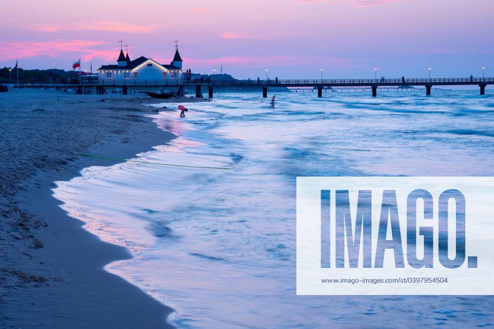 Quiet Abendstimmung on Beach with quiet Waves and Two Person near a Seabridge with Building in the Dusk, clearer, Blue purple Sky with Pink Clouds, Seabridge Ahlbeck at last Evening light, Motion blur, Wiping effect, Long exposure, View from Western Beach, Seaside resort Ahlbeck, Island Usedom, Mecklenburg Western Pomerania, Germany, Europe
