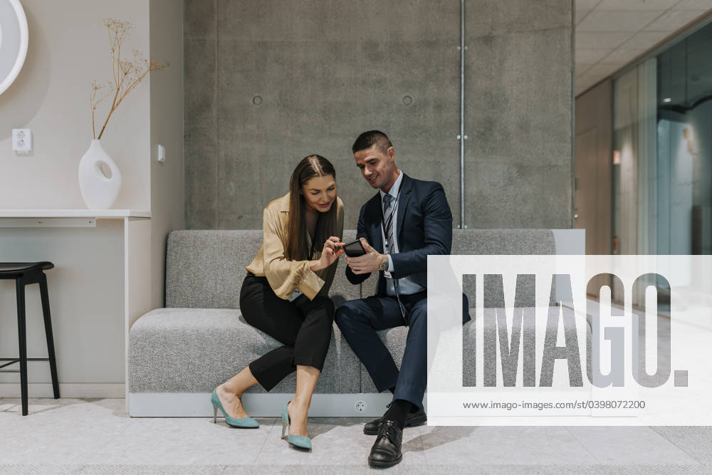 Full length of male and female delegates sitting on sofa sharing smart phone during seminar Sweden, Gothenburg, model released, property released