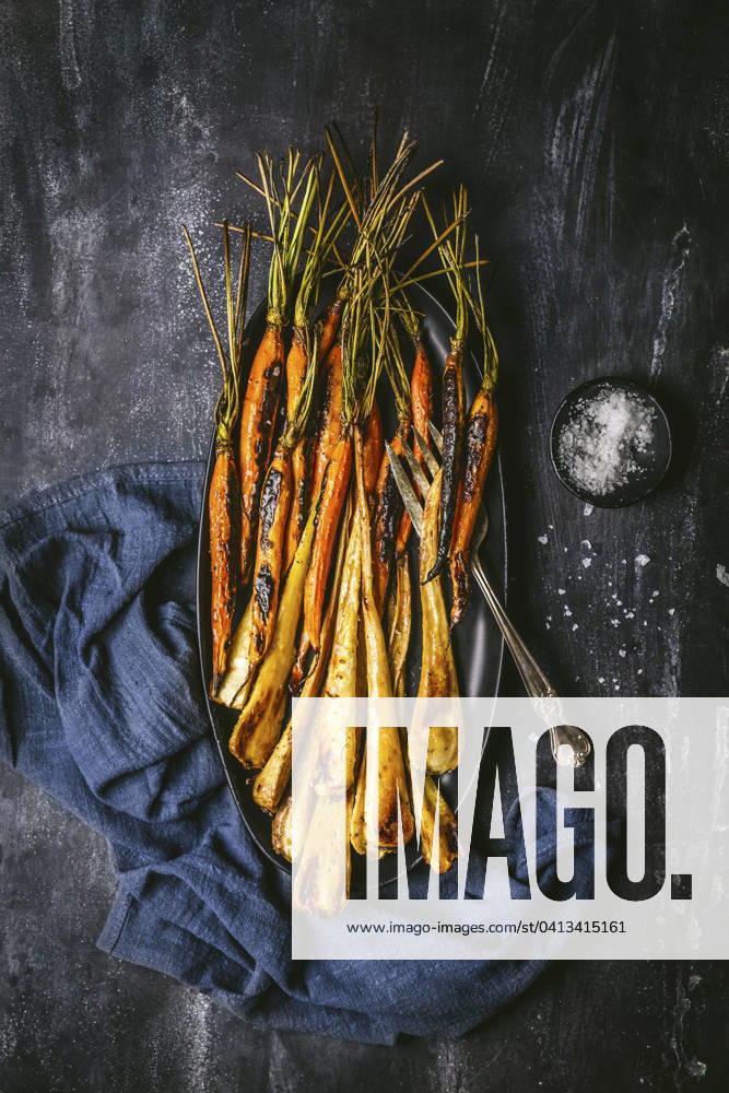 Roasted, glazed carrots and parsnips arranged on a dark oval platter with serving fork, and blue linen napkin on dark background Roasted, glazed carrots and parsnips arranged on a dark oval platter with serving fork, and blue linen napkin on dark background