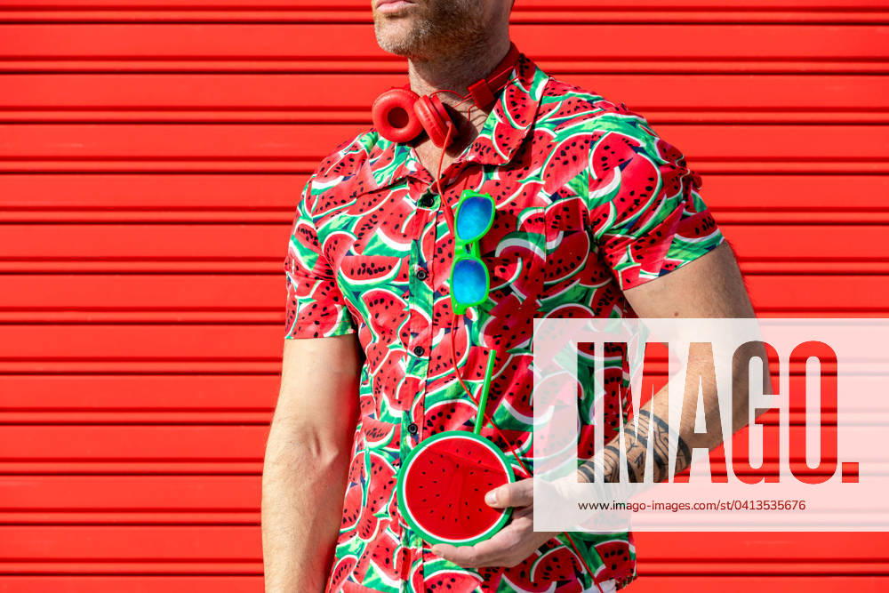 Crop unrecognizable male looking away while standing wearing printed water melon slices shirt and carrying mock juice with straw in hand against red background Model Released SergioVictorVega_ID19622_581836_001