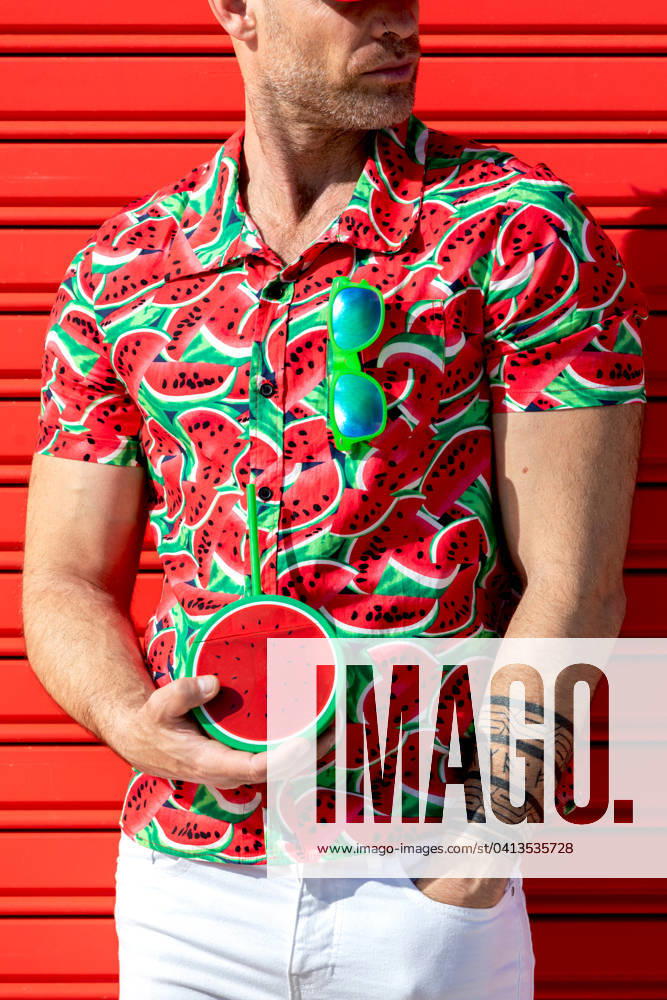 Crop unrecognizable male looking away while standing wearing printed water melon slices shirt and carrying mock juice with straw in hand against red background Model Released SergioVictorVega_ID19622_581838_003