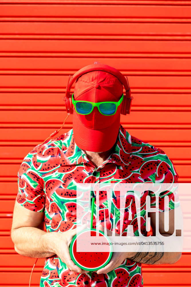 Anonymous man in printed shirt covering face with hat Unrecognizable street style male with hat covering face and wearing sunglasses headphones in place while standing in printed water melon slices shirt and carrying mock juice against red background Model Released SergioVictorVega_ID19622_581839_004