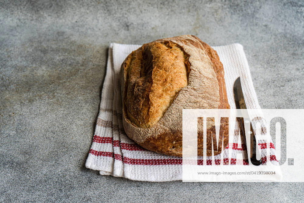Top view of golden-brown rye sourdough bread on a white cloth with red stripes, next to a bread knife on a textured surface Model Released AnnaBogush_ID19733_585087_007