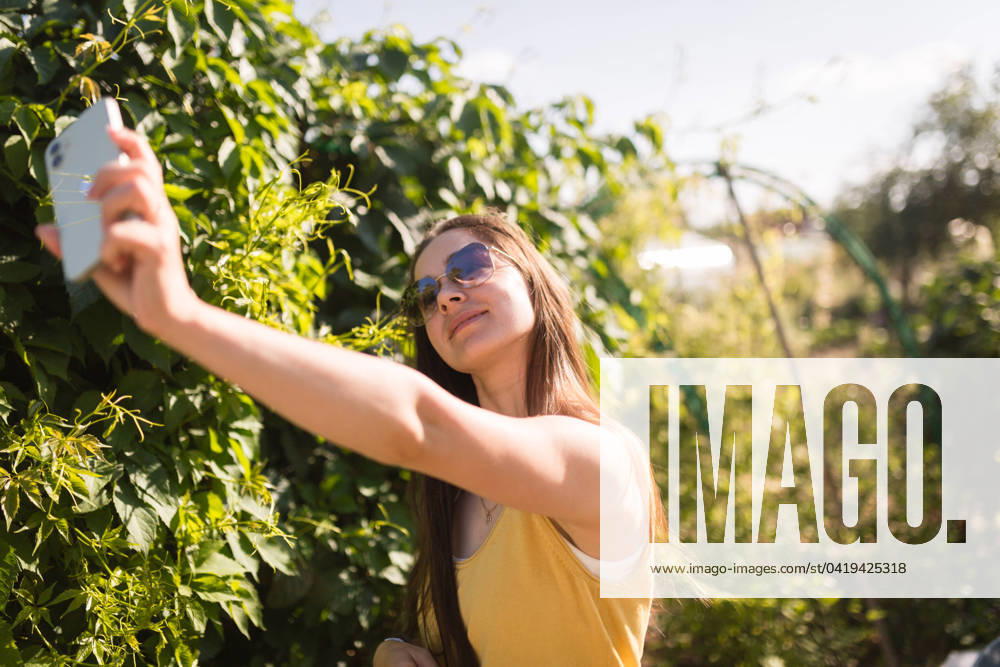IMAGO Creative: Sommer, Sommertage im Garten MR_ID19530_16353_MarinaGabitova.jpg A cheerful young woman in a yellow tank top takes a selfie with her phone among lush greenery on a sunny day Model Released Declaria_ID19530_579187_001