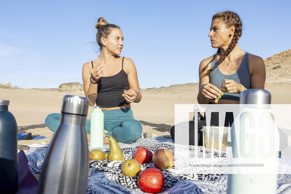 Two women enjoying a nutritious picnic on a beach mat, surrounded by fresh fruit and water bottles, engaged in a conversation with a clear blue sky above. Model Released JuanAizpuru_ID19654_582744_024