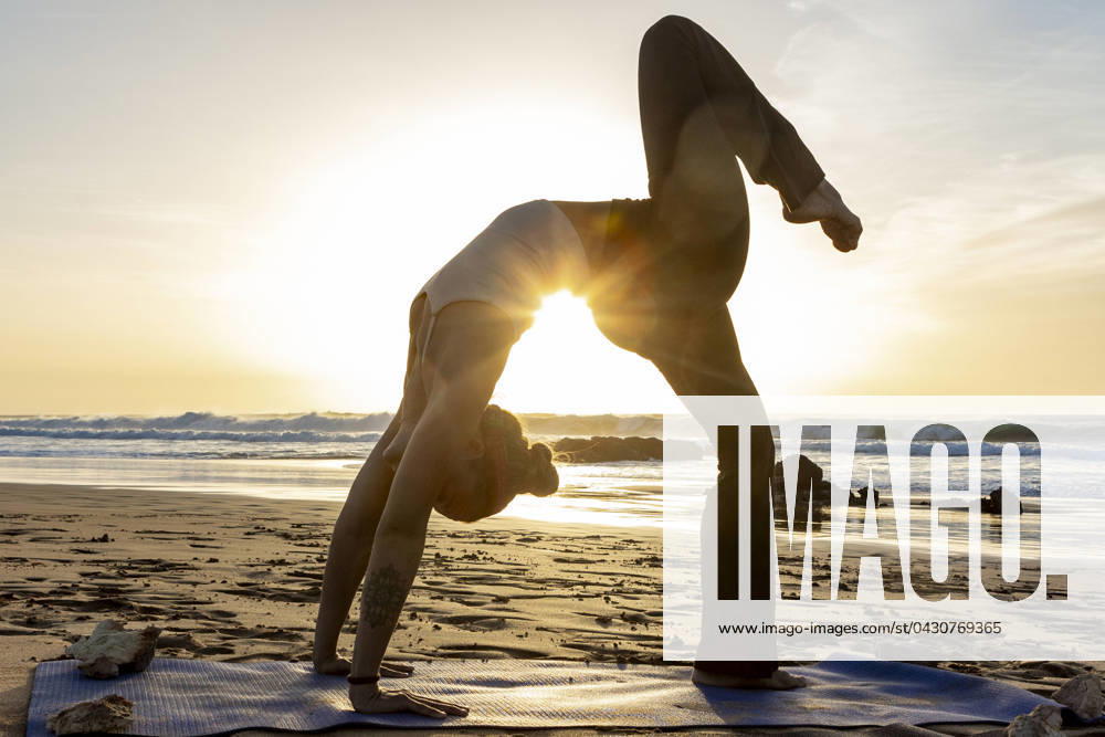 A yoga feamle practitioner performs a challenging pose on a sandy beach as the sun sets, creating a tranquil and inspiring setting. Model Released JuanAizpuru_ID19654_582782_040
