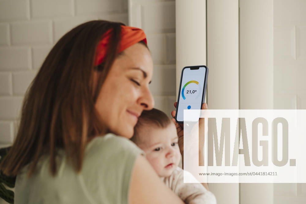IMAGO Creative, Jahreszeit Sommer, Hitze Mother embracing baby daughter and adjusting radiator through smart phone at home model released, Symbolfoto property released, EBBF08730