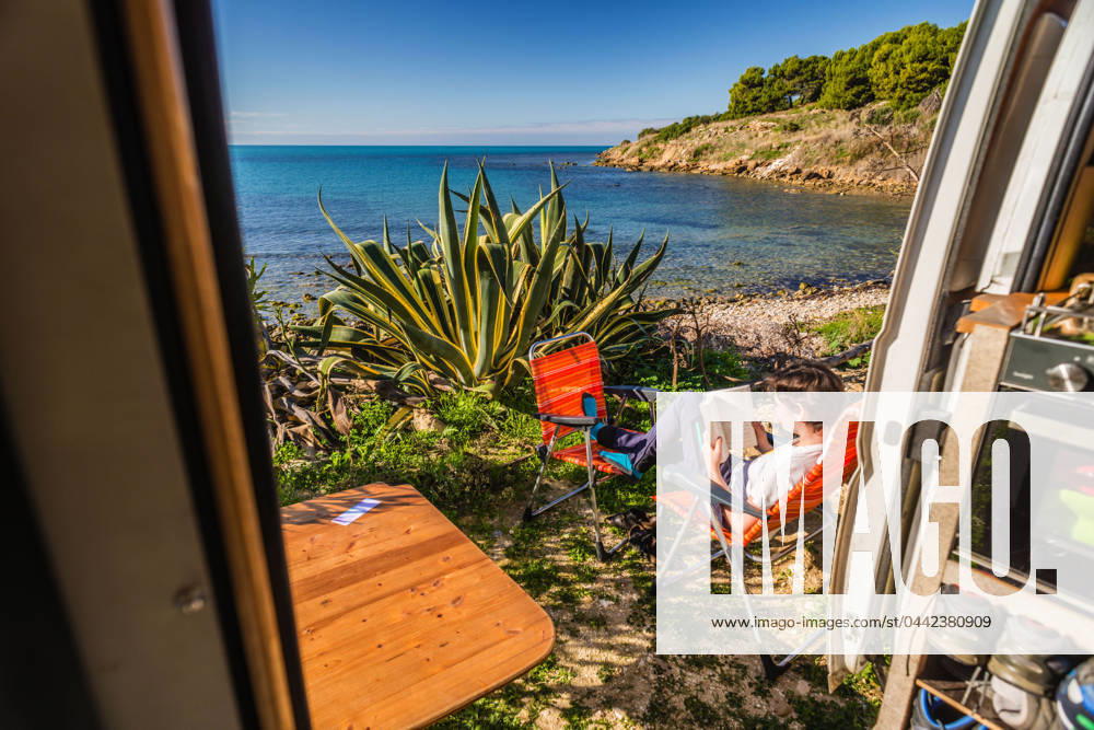 View from the sliding door of a motorhome standing on a pitch on the unpaved shore on the Mediterranean coast, a young woman sits on camping chairs and reads a book Sicily, Italy A motorhome stands on a pitch on the shore of the Mediterranean Sea