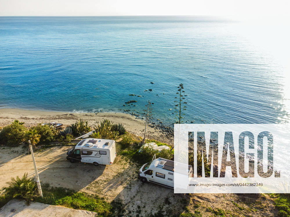 A camper van and an alcove motorhome are parked on a pitch on the unpaved shore on the Mediterranean coast, boats lie in front of them on the beach Sicily, Italy A motorhome is parked on a pitch on the shore of the Mediterranean Sea