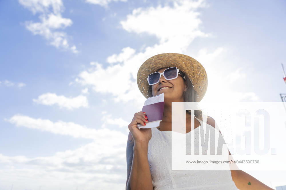 Cheerful woman in straw hat and sunglasses holding a passport, ready for a leisure trip under a sunny sky, symbolizing a carefree weekend getaway. Model Released JuanAizpuru_ID20118_595637_034