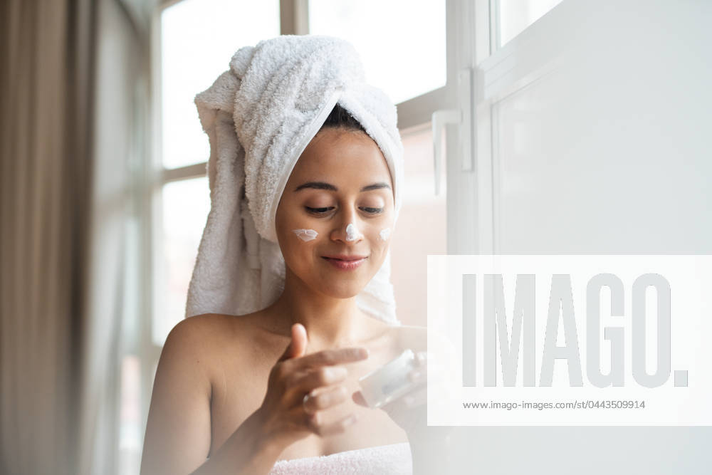 A woman with a towel-wrapped hair applies facial moisturizer after a shower, reflecting a daily beauty regimen Model Released PabloMenendez_ID20332_601469_056