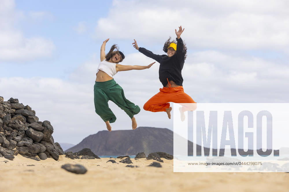 A joyful same-sex couple leaps into the air on a sandy beach, with a scenic mountain backdrop, embodying freedom and happiness Model Released JuanAizpuru_ID20478_604918_019