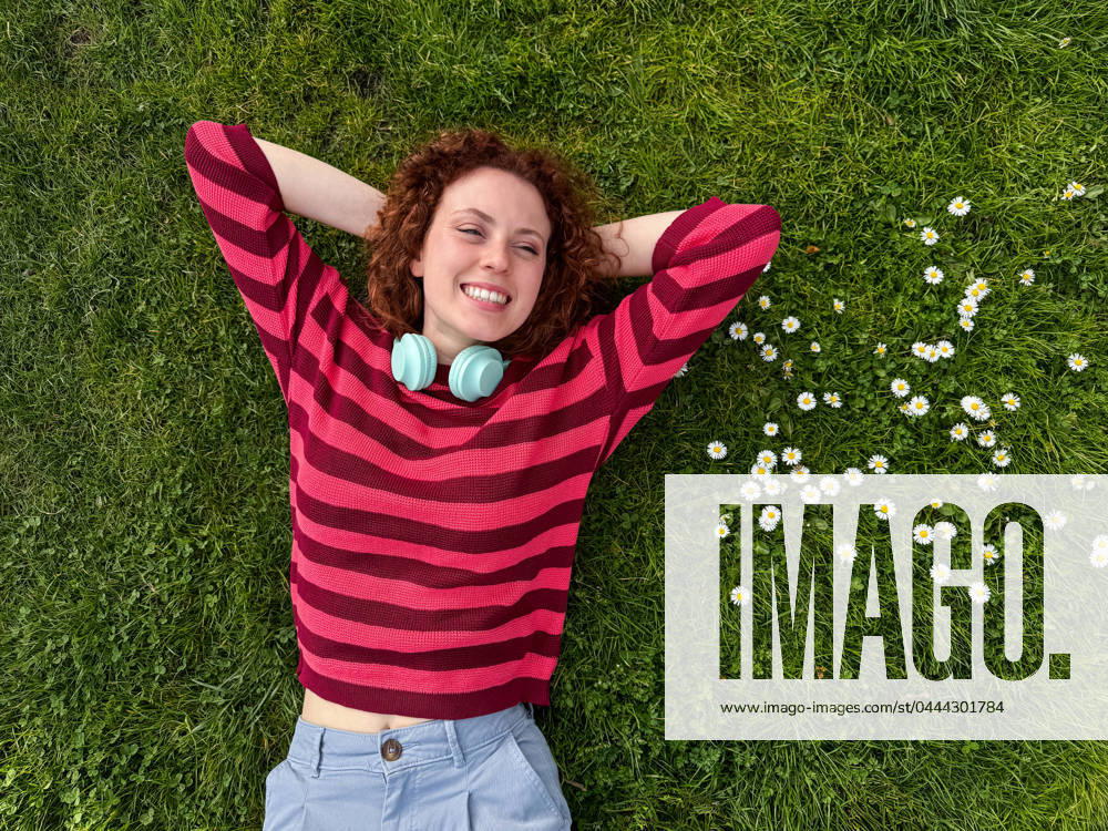 IMAGO Creative: Sommer, Sommertage im Stadtpark A happy young redhead woman with curly red hair lies on a lush green lawn, surrounded by white daisies, enjoying a leisurely day Model Released ManuelRuiz_ID20916_617113_046