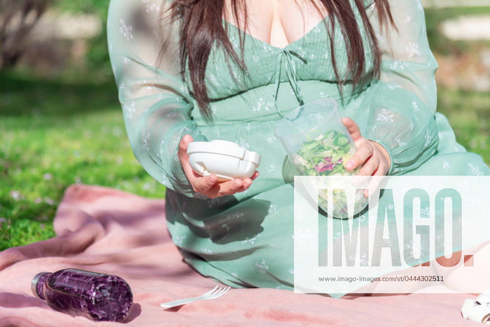 IMAGO Creative: Sommer, Sommertage im Stadtpark A woman sits on a picnic blanket, holding a bowl of fresh salad and a set of white earbuds, enjoying the sunshine. Model Released NataliaPenado_ID20535_606437_012