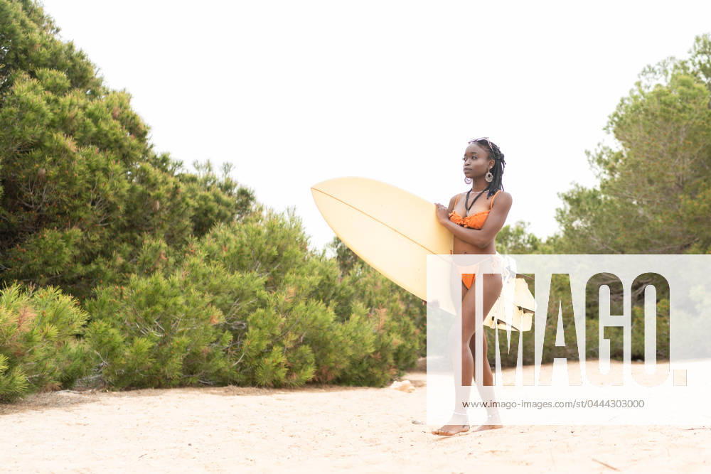 An African American girl in an orange bikini holds a surfboard at the beach, surrounded by natural vegetation Model Released OscarDeDios_ID20936_617642_003