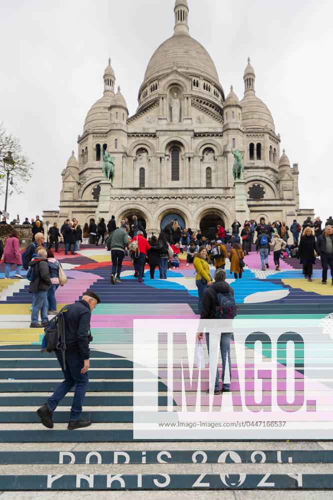 Paris, France April 19, 2024 - People walk up the stairs leading to the Sacre Coeur basilica decorated with a fresco in Olympic colors as part of the preparations for the Paris 2024 Olympic Games. Less than 100 days remain for the Olympic and Paralympic Games organizers to finalize preparations for the event that will welcome millions of visitors from July 16. OLY, JEUX OLYMPIQUES PARIS 2024, DECORATIONS, FRESQUE, ILLUSTRATION, MONTMARTRE, BASILIQUE DU SACRE COEUR, SPORT, ESCALIERS, MARCHES COLOREES,
