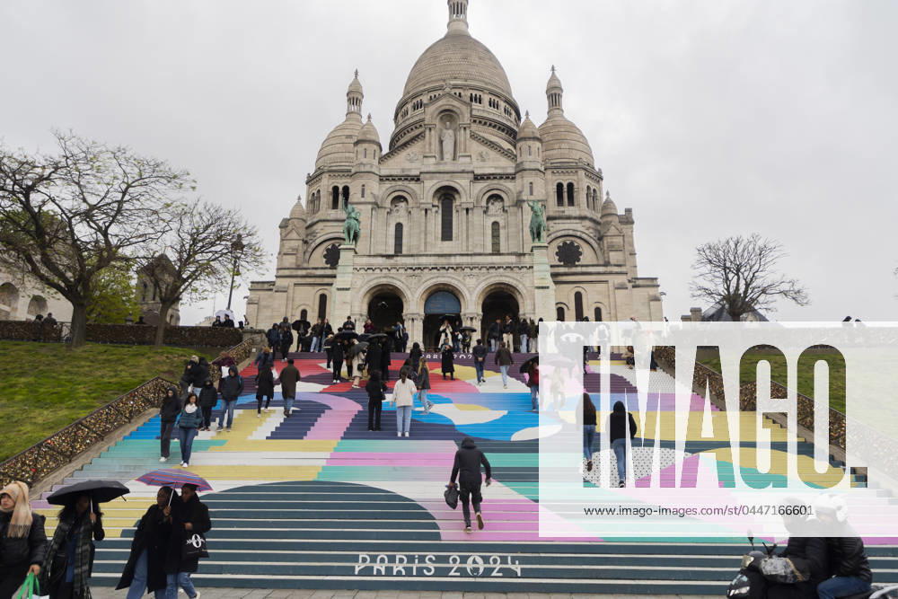 Paris, France April 19, 2024 - People walk up the stairs leading to the Sacre Coeur basilica decorated with a fresco in Olympic colors as part of the preparations for the Paris 2024 Olympic Games. Less than 100 days remain for the Olympic and Paralympic Games organizers to finalize preparations for the event that will welcome millions of visitors from July 16. OLY, JEUX OLYMPIQUES PARIS 2024, DECORATIONS, FRESQUE, ILLUSTRATION, MONTMARTRE, BASILIQUE DU SACRE COEUR, SPORT, ESCALIERS, MARCHES COLOREES,