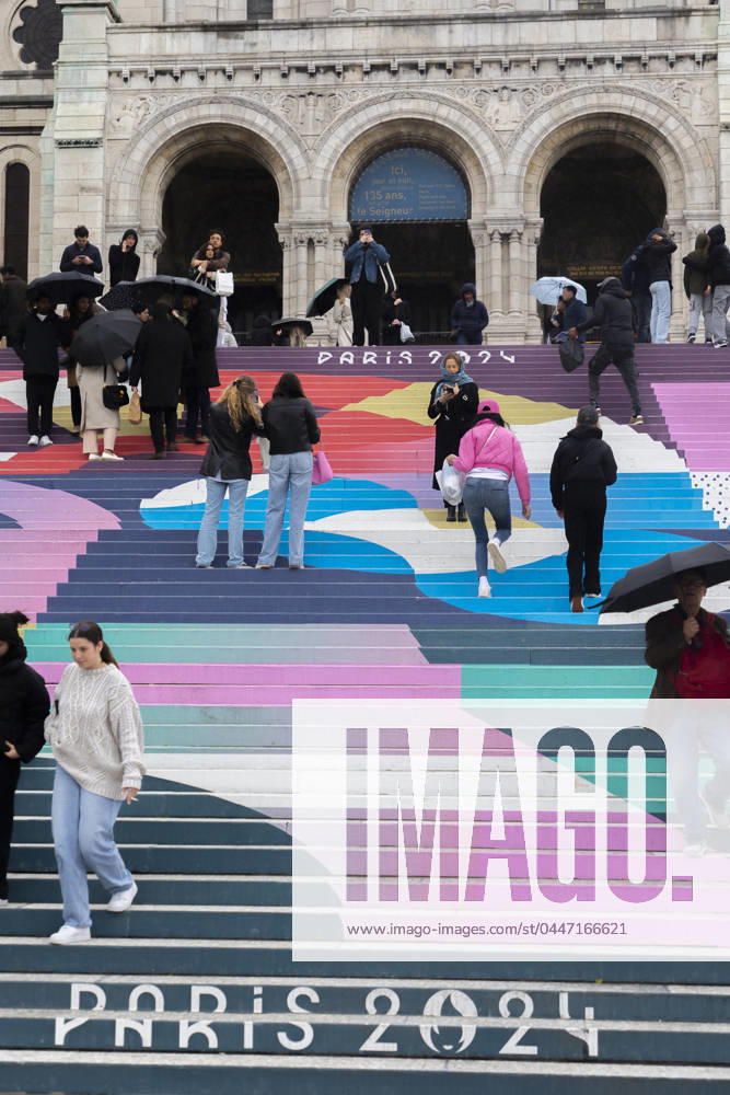 Paris, France April 19, 2024 - People walk up the stairs leading to the Sacre Coeur basilica decorated with a fresco in Olympic colors as part of the preparations for the Paris 2024 Olympic Games. Less than 100 days remain for the Olympic and Paralympic Games organizers to finalize preparations for the event that will welcome millions of visitors from July 16. OLY, JEUX OLYMPIQUES PARIS 2024, DECORATIONS, FRESQUE, ILLUSTRATION, MONTMARTRE, BASILIQUE DU SACRE COEUR, SPORT, ESCALIERS, MARCHES COLOREES,