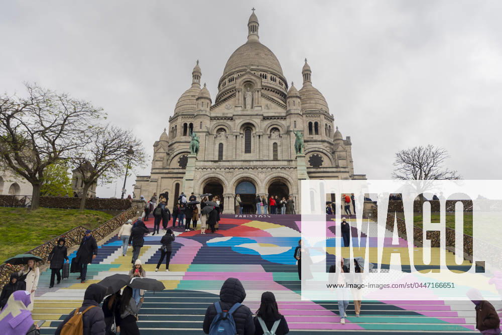 Paris, France April 19, 2024 - People walk up the stairs leading to the Sacre Coeur basilica decorated with a fresco in Olympic colors as part of the preparations for the Paris 2024 Olympic Games. Less than 100 days remain for the Olympic and Paralympic Games organizers to finalize preparations for the event that will welcome millions of visitors from July 16. OLY, JEUX OLYMPIQUES PARIS 2024, DECORATIONS, FRESQUE, ILLUSTRATION, MONTMARTRE, BASILIQUE DU SACRE COEUR, SPORT, ESCALIERS, MARCHES COLOREES,
