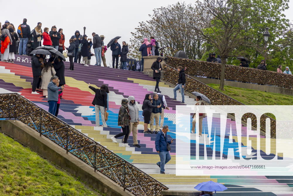 Paris, France April 19, 2024 - People walk up the stairs leading to the Sacre Coeur basilica decorated with a fresco in Olympic colors as part of the preparations for the Paris 2024 Olympic Games. Less than 100 days remain for the Olympic and Paralympic Games organizers to finalize preparations for the event that will welcome millions of visitors from July 16. OLY, JEUX OLYMPIQUES PARIS 2024, DECORATIONS, FRESQUE, ILLUSTRATION, MONTMARTRE, BASILIQUE DU SACRE COEUR, SPORT, ESCALIERS, MARCHES COLOREES,