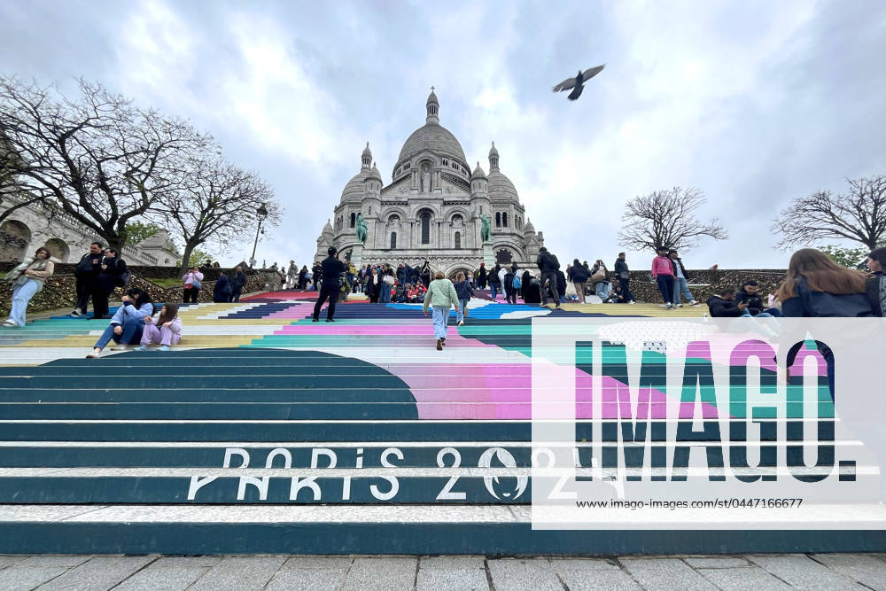 Paris, France April 19, 2024 - People walk up the stairs leading to the Sacre Coeur basilica decorated with a fresco in Olympic colors as part of the preparations for the Paris 2024 Olympic Games. Less than 100 days remain for the Olympic and Paralympic Games organizers to finalize preparations for the event that will welcome millions of visitors from July 16. OLY, JEUX OLYMPIQUES PARIS 2024, DECORATIONS, FRESQUE, ILLUSTRATION, MONTMARTRE, BASILIQUE DU SACRE COEUR, SPORT, ESCALIERS, MARCHES COLOREES,