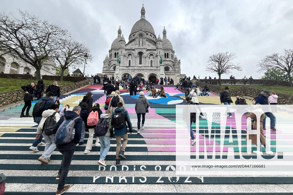 Paris, France April 19, 2024 - People walk up the stairs leading to the Sacre Coeur basilica decorated with a fresco in Olympic colors as part of the preparations for the Paris 2024 Olympic Games. Less than 100 days remain for the Olympic and Paralympic Games organizers to finalize preparations for the event that will welcome millions of visitors from July 16. OLY, JEUX OLYMPIQUES PARIS 2024, DECORATIONS, FRESQUE, ILLUSTRATION, MONTMARTRE, BASILIQUE DU SACRE COEUR, SPORT, ESCALIERS, MARCHES COLOREES,