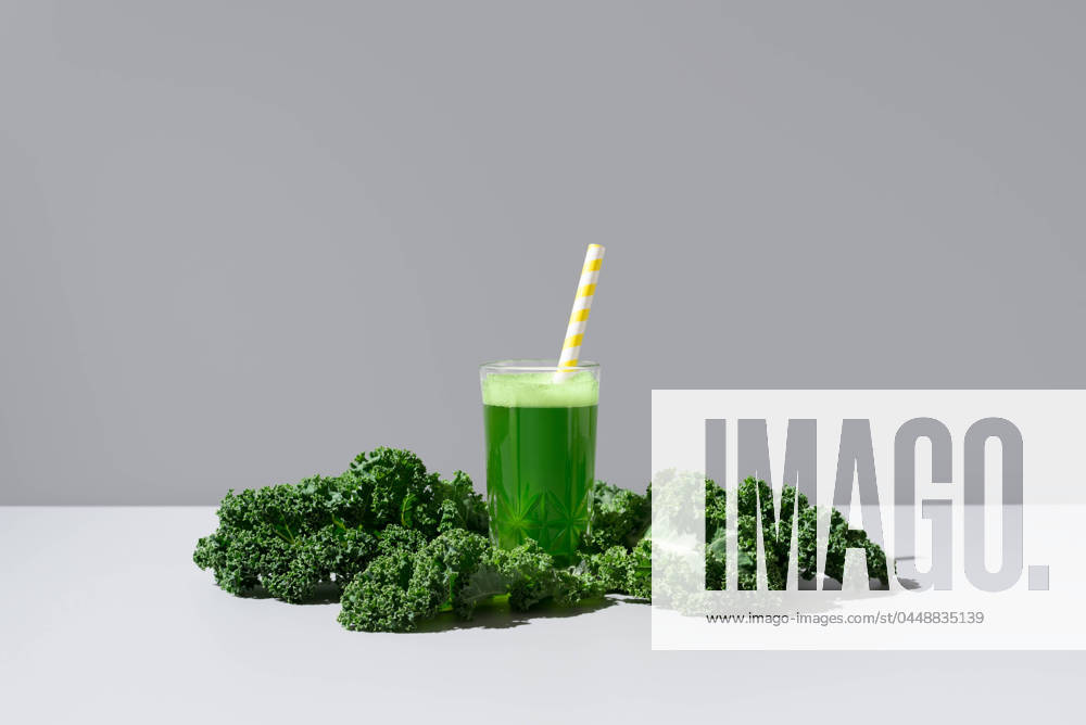 A vibrant green kale juice served in a transparent glass with a yellow-striped straw, accompanied by fresh kale leaves on a neutral background Model Released RuthCarrodeguas_ID21186_624726_006