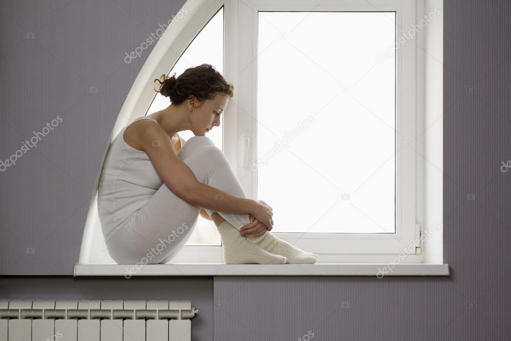 Mid adult woman sits on windowsill with ankles crossed model released