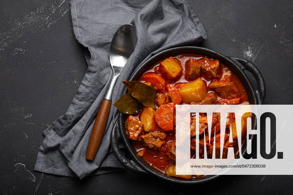 IMAGO Creative, Herbst, Food, Gulasch Beef meat stew with potatoes, carrot and delicious gravy in black casserole pot with bay leaves with spoon on black dark rustic concrete background from above . Beef meat stew with potatoes, carrot and delicious gravy in black casserole pot with bay leaves with spoon on black dark rustic concrete background from above .