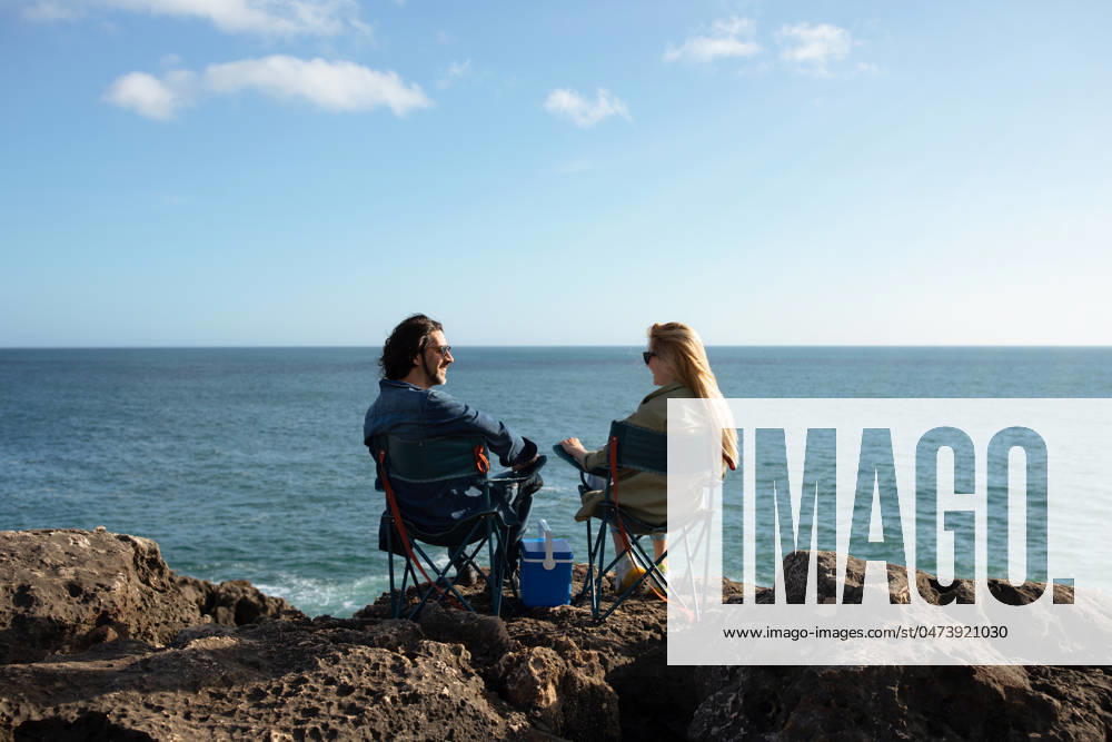 Happy Young Couple Sitting In Camping Chairs On Beach Rocks Near Ocean Happy Young Couple Sitting In Camping Chairs On Beach Rocks Near Ocean, Smiling Man And Woman Having Picnic Or Outdoor Date, Millennial Lovers Chatting And Looking At Water, Enjoying View, Copy Space