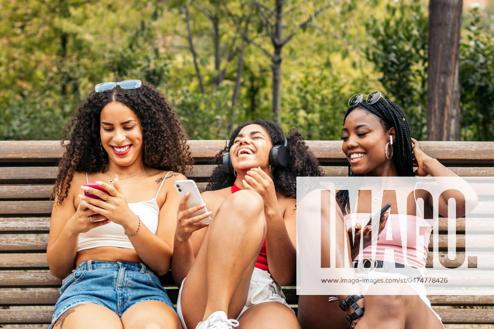 IMAGO Creative: Sommer, Sommertage im Stadtpark African american female friends sitting on a park bench laughing and having fun while using smartphone and listening to music with headphones. Model Released PedroMerino_ID18922_563471_005