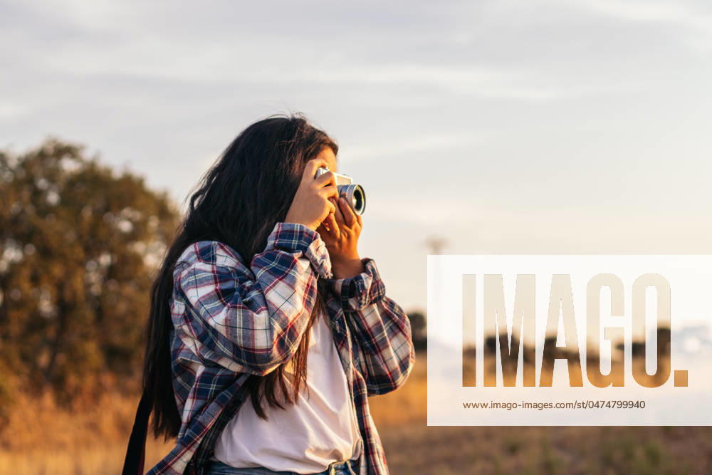 Young woman in checkered shirt and casual clothes taking photos with an analog camera in the field at sunset. The image conveys the concept of retro and vintage Model Released PedroMerino_ID19010_565582_006