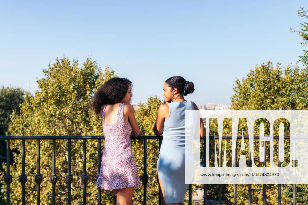 IMAGO Creative: Sommer, Sommertage im Stadtpark Back view of two African-American girls in dresses, looking at the city views and a large urban park while leaning on a bridge railing. They talk and enjoy the day together. Model Released PedroMerino_ID19012_565710_023