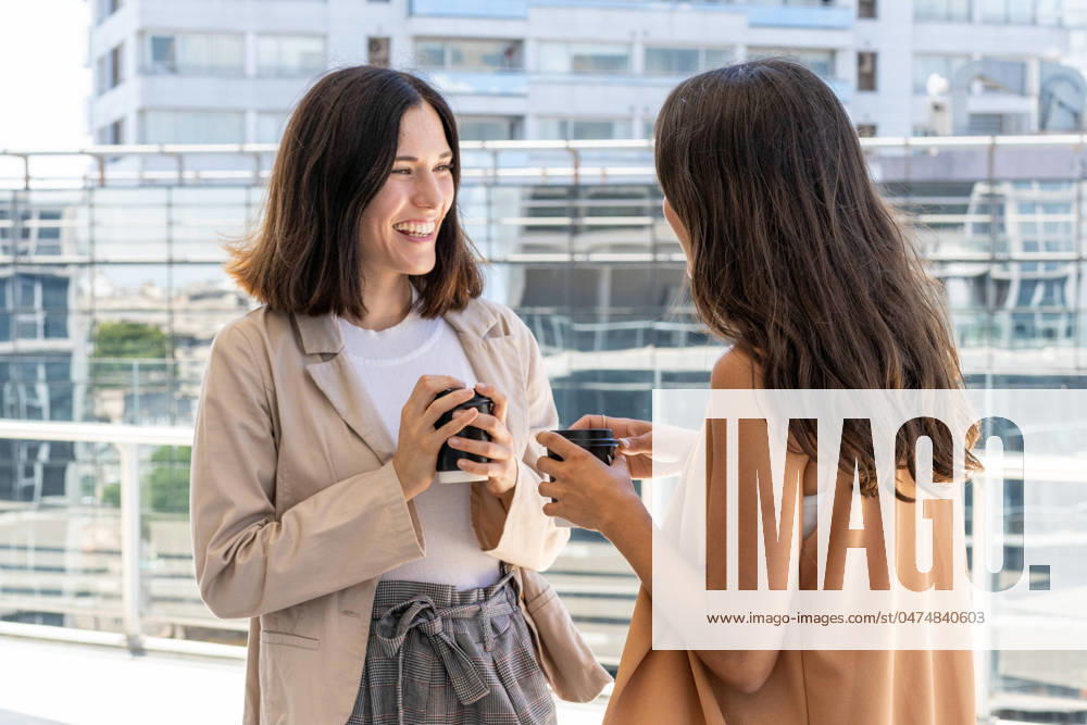 Two businesswomen are standing outside a building, enjoying their office break. They are smiling and holding cups of coffee, indicating a sense of camaraderie and friendship. Model Released ValeriaVenezia_ID20934_617621_025