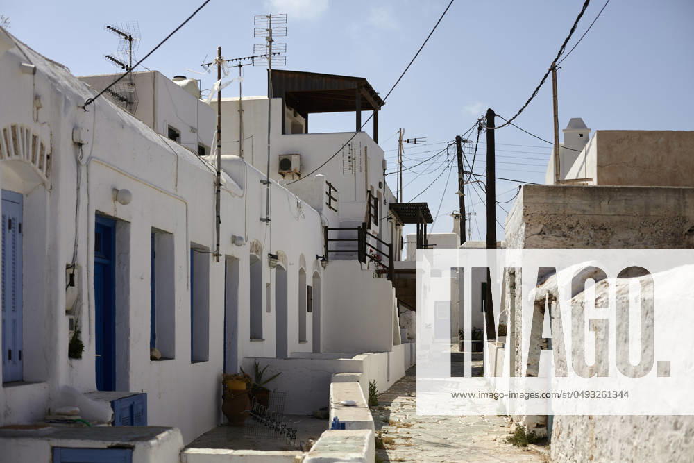 A narrow alley in a traditional Mediterranean village with white buildings and blue doors under a clear sky, with electrical wires crisscrossing overhead. Folegandros Greece bola01001